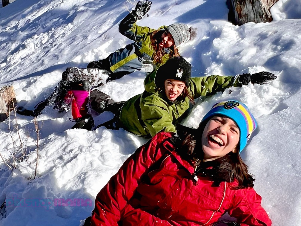 Girls in multi-color jackets and pants laughing and rolling in the snow while taking family snow pictures