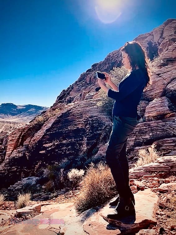 Pix Snap Mama in dark blue top, dark gray pants, and black boots standing on a rock in the desert Southwest taking a picture with her iPhone.