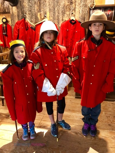 Three girls dressed up as Canadian Mounties with red military jackets and hats. On the left has a yellow hat and sword, in the middle has a white safari style hat, white gloves and a riding crop, on the right has a brown wide brim mountie hat.