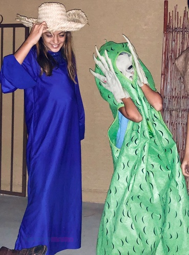 Two girls dressed up as "aliens". On left is in blue long sleeve gown with pillow stomach and straw farmer hat. On right has green pickle costume with green rubber alien mask and hands.