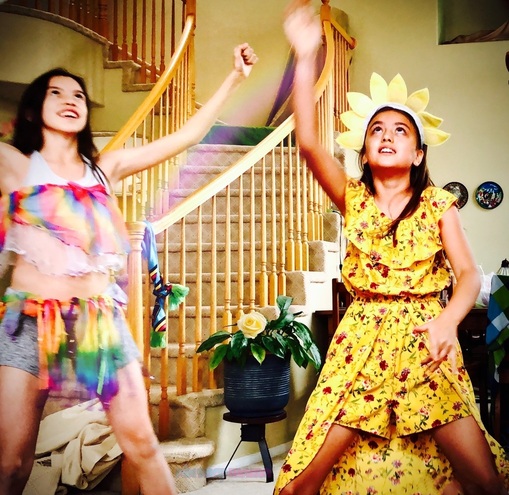 Two girls play dress up. On the left has rainbow tassels as a top and skirt. On the right has a yellow skort jumper with red flower pattern and a yellow sunflower headband.