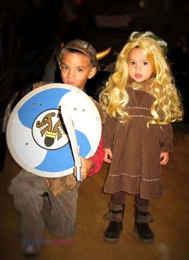 Two little kids dress up like Vikings. The boy has a shield, sword, and helmet. The girl has brown dress and long blond wig with braids & moss decoration.