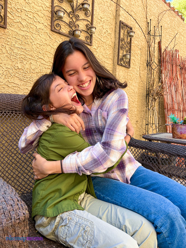 iPhone portraits of two laughing girls sitting and hugging on an outdoor sofa. Pix Snap Mama is overlaid in the lower left corner