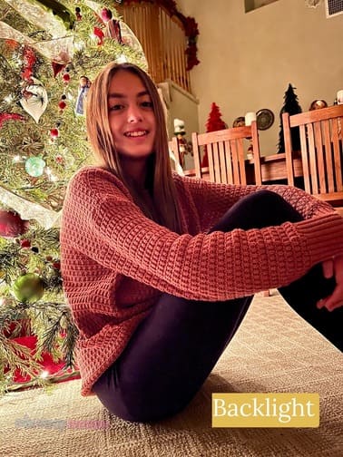 A girl with light hair wearing a red long sleeve top and dark pants sitting with her arms around her knees in front of a Christmas tree. A backlight example of the different types of photography lighting.