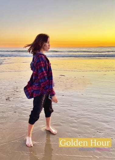 A girl wearing a purple and blue button down and rolled up dark pants walking along the beach during a beautiful yellow and orange hued sunset. A golden hour light example of the different types of photography lighting.