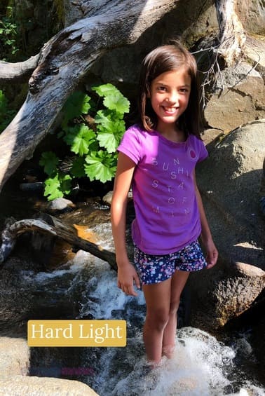 A young girl in purple top and blue shorts standing in a stream in front of rocks and a fallen branch. A hard light example of the different types of photography lighting.