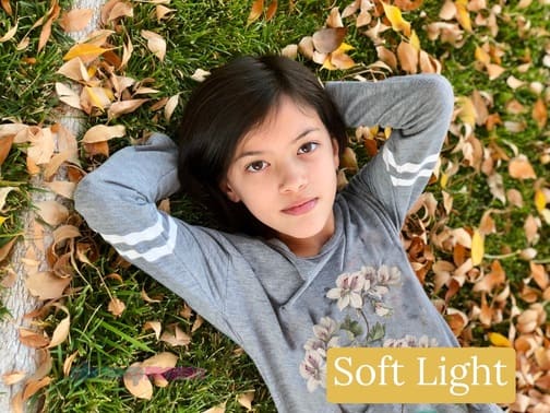 A young girl with dark hair wearing a light gray long sleeve shirt laying in the grass and leaves under a tree. A soft light example of the different types of photography lighting.